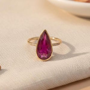 May include: A gold ring with a large, teardrop-shaped, deep magenta gemstone. The gold band has small, clear accent stones. The ring is set against a neutral, textured background, highlighting the vibrant colour of the gemstone.