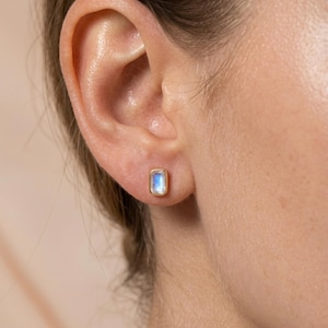 Puede incluir: Primer plano de un pendiente de oro con una piedra preciosa rectangular, iridiscente, azul claro. El pendiente está engastado en un bisel de oro y se lleva en una oreja. El fondo es de un color suave y neutro.