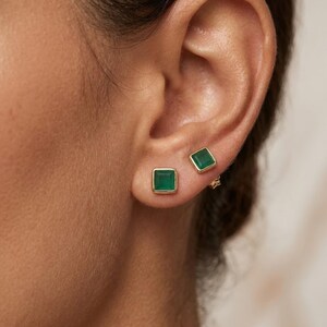 Puede incluir: Un par de pendientes de botón de forma cuadrada con piedras de color verde vibrante engastadas en metal pulido de tono dorado. Los pendientes se llevan en el lóbulo de la oreja, mostrando una superficie lisa y plana con un diseño geométrico.