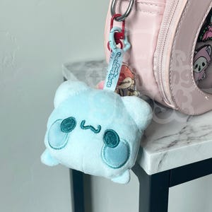 May include: A light blue plush bear keychain with embroidered details, including a smiling face and rounded features. The keychain is attached to a pink bag with a clear front displaying various pins. The keychain reads "Bonding Blossoms".