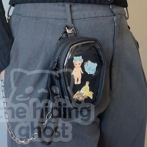 Puede incluir: Bolso bandolera negro con un panel frontal transparente, adornado con pegatinas de dibujos animados de colores. El bolso está sujeto a una cadena plateada y se lleva en la cintura. El bolso es rectangular y tiene cierre de cremallera.