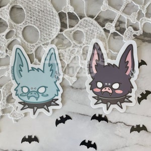 May include: Two cute cartoon bat stickers with punk rock collars. The bat on the left is light blue with a light blue collar and the bat on the right is dark purple with a black collar.