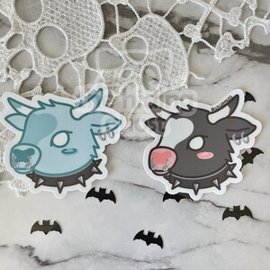 May include: Two cartoon cow stickers with black studded collars and silver rings in their noses and ears. The cow on the left is light blue with white spots and the cow on the right is black with white spots. Both cows have pink noses.