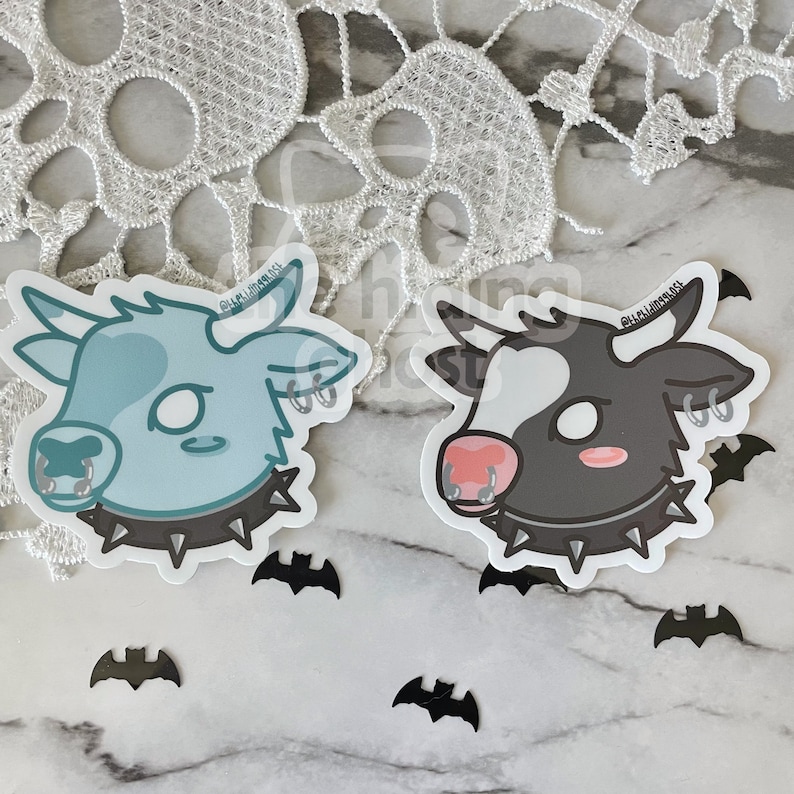 May include: Two cartoon cow stickers with black studded collars and silver rings in their noses and ears. The cow on the left is light blue with white spots and the cow on the right is black with white spots. Both cows have pink noses.