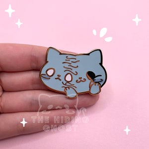 May include: A light blue cat-shaped enamel pin with a gold border. The pin features a stylized cat design with a closed eye and the text "THE HIDING GHOST" at the bottom. The pin is held in a hand against a pink background.