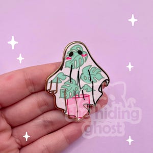 May include: A gold-colored enamel pin featuring a white ghost holding a potted plant with green leaves. The ghost has a pink blush and black eyes. The pin is on a hand.