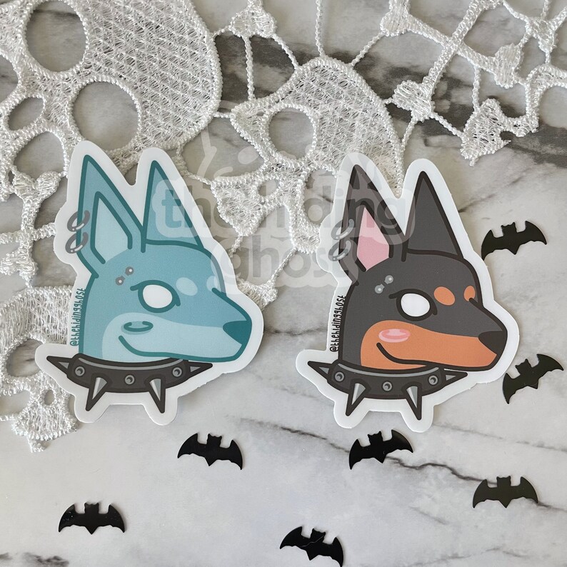 May include: Two sticker designs of dogs with punk rock accessories. The dog on the left is light blue with a silver nose ring and a studded collar. The dog on the right is black and brown with a silver nose ring and a studded collar. Both dogs have a white eye and a pink cheek.