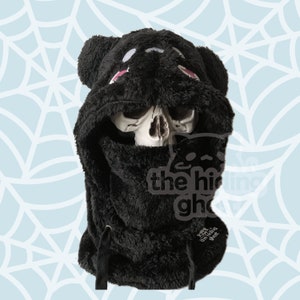 May include: Black hooded fleece with a bear face and pink accents. The hood is pulled up and covers the face. The text "the hiding ghost" is visible on the front of the hood.