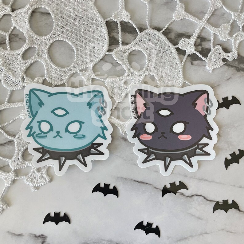 May include: Two sticker designs of cartoon cats with a third eye and a spiked collar. The cat on the left is light blue and the cat on the right is dark purple.