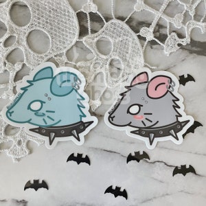 May include: Two cartoon rat stickers with punk rock accessories. The rat on the left is light blue with a black studded collar. The rat on the right is grey with a pink ear and a black studded collar. Both rats have black eyes and white pupils. The stickers are on a white background with black bats.