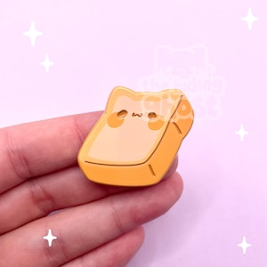 May include: A yellow and orange enamel pin in the shape of a cat-shaped piece of toast. The pin has a cute face with small eyes and a mouth. The pin is held in a hand against a pink background with white star accents.