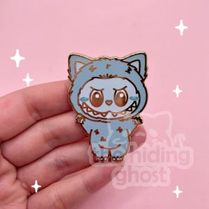 Puede incluir: Pin esmaltado azul claro con un personaje de dibujos animados disfrazado de monstruo. El pin tiene un contorno dorado y una cara sonriente con dientes afilados. El personaje lleva una capucha con orejas de gato. Fondo rosa.