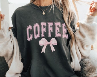 May include: A dark gray t-shirt with the word "COFFEE" printed in pink letters with a pink bow underneath. The shirt is being worn by a person.