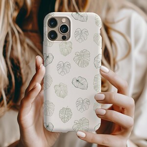 May include: White phone case with a pattern of green leaves. The case has a camera lens cutout and a white background.