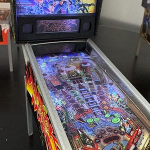 May include: A small pinball machine with the words "Attack from Mars". The backboard has a colourful graphic of aliens and a spaceship. The playfield is detailed with various targets and bumpers. The machine is lit with blue lights.