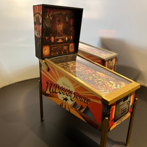 Könnte beinhalten: Eine Miniatur-Nachbildung des Indiana Jones Flippers. Die Maschine ist aus Holz und Metall und verfügt über eine gläserne Spielfläche. Die Maschine ist in einem rot-orange-goldenen Farbschema lackiert und trägt den Text "Indiana Jones: Der Tempel des Schicksals".