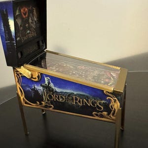 Può includere: Mini flipper da tavolo a tema "Il Signore degli Anelli". Il mobile è blu e oro, con il logo e le immagini del film. Ha un piano di gioco in vetro trasparente e gambe dorate. Un oggetto da collezione.