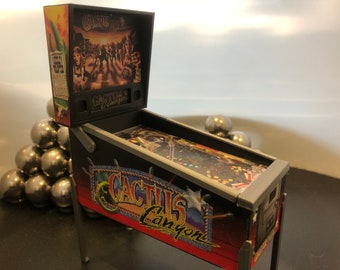 May include: A small-scale pinball machine with a "Cactus Canyon" design. The cabinet is black with red and yellow details. The playfield has intricate graphics, and the backboard depicts a desert scene. Silver metal balls are also visible.
