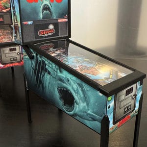 Könnte beinhalten: Miniatur-Flipperautomat mit dem Thema "Der weiße Hai". Die Maschine hat einen schwarzen Rahmen und Beine, mit Grafiken eines Hais und dem Filmtitel. Die Rückwand zeigt "JAWS" in roten Buchstaben. Ein Sammlerstück.