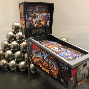May include: A miniature pinball machine with a silver and black cabinet. The playfield features a colourful design with the words "Junk Yard" printed on the side. The machine is sitting on a table with a pile of silver balls in the foreground.