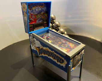 May include: A miniature pinball machine with a blue and silver cabinet. The game is called "Whirlwind" and features a colourful, detailed playfield.