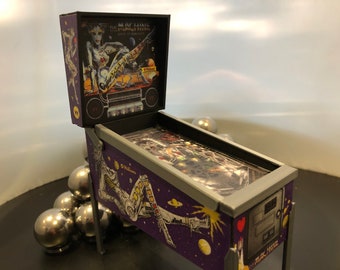 May include: A miniature pinball machine with a purple and silver design. The backglass features a woman in a futuristic outfit with the text "The Machine - The Best of Journey".