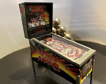May include: A miniature pinball machine with a black and silver cabinet. The game is called "Attack from Mars" and features a colorful, detailed playfield with a red, yellow, and black theme. The machine is open, revealing the playfield and the backglass with a graphic of a spaceship and aliens.