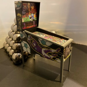 May include: A miniature pinball machine with a black and white soccer theme. The machine has a black cabinet with a clear glass playing field. The backglass features a soccer player in a purple jersey and a soccer ball. The machine is on a black surface.