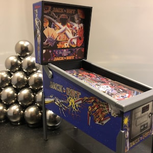 May include: A miniature pinball machine with a blue and silver design. The machine is titled "Jack Bot" and features a graphic of a robotic hand reaching out to another hand. The backglass features a colorful image of a woman and a robot.