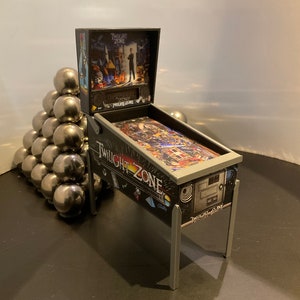 May include: A miniature pinball machine with a black and silver design. The machine features a "Twilight Zone" theme with a black and white image of a man in a suit on the back glass. The machine is on a black surface.