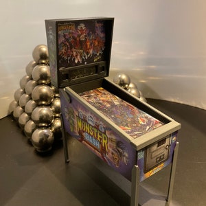 May include: A miniature pinball machine with a gray metal frame and a purple, blue, and green playfield. The playfield features the text "Monster Bash" and a graphic of a monster with a green face. The machine is sitting on a black surface.