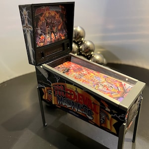 May include: A miniature pinball machine with a black and silver cabinet. The game is called "Ye Olde Medieval Madness". The cabinet has a glass top and a raised silver trim. The backglass features a medieval scene with a sword and a knight.