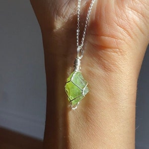 May include: A silver necklace with a light green crystal pendant. A beaded bracelet with various colored beads is also visible. The jewelry is displayed on a person's arm.