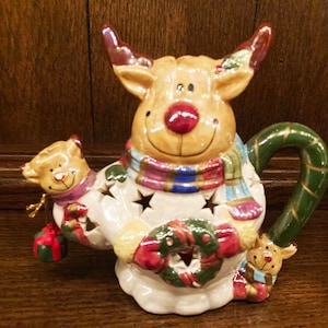 May include: A ceramic reindeer candle holder with a green and gold handle. The reindeer is wearing a colorful scarf and holding a wreath. There are two smaller reindeer figures on either side of the main reindeer.