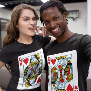 May include: Two black crew neck t-shirts featuring large graphic prints of playing cards. One shirt displays the Queen of Hearts with a red heart symbol and the letter Q, while the other shirt features the King of Hearts with a red heart and the letter K.