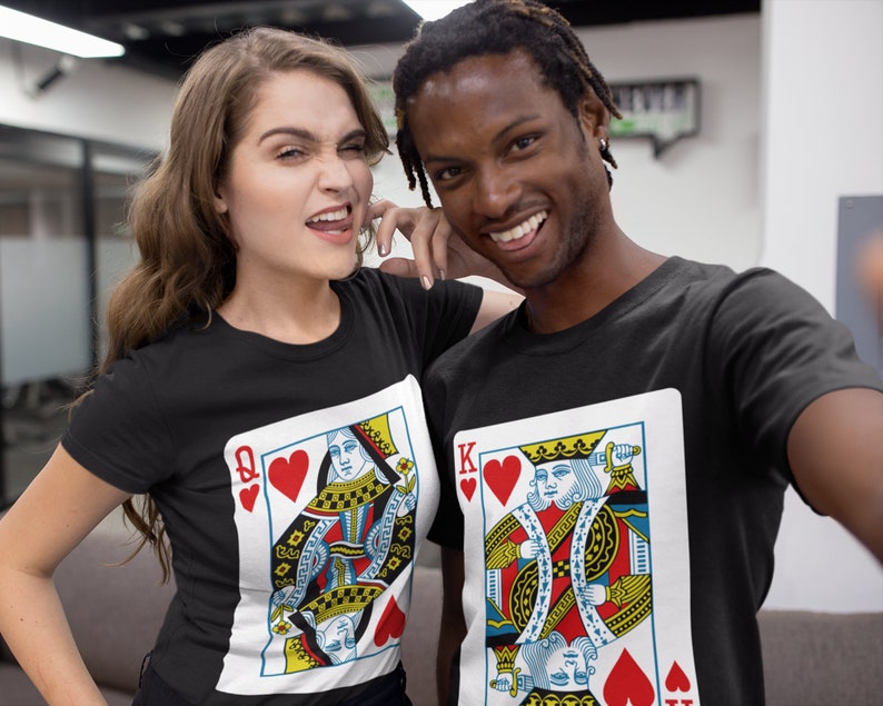 May include: Two black crew neck t-shirts featuring large graphic prints of playing cards. One shirt displays the Queen of Hearts with a red heart symbol and the letter Q, while the other shirt features the King of Hearts with a red heart and the letter K.