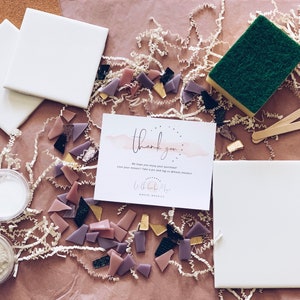 May include: A mosaic tile craft kit with two white ceramic tiles, a small container of white grout, a small container of gray grout, a sponge, two wooden sticks, and a variety of small, colorful glass tiles. The kit also includes a thank you card with the text "Thank you. We hope you enjoy your purchase! Love your mosaic? Tag us on social media! #LittleBits #MosaicTiles".