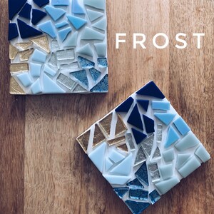 Puede incluir: Dos posavasos cuadrados con un dise&ntilde;o de mosaico en tonos azules, dorados y plateados. Los posavasos est&aacute;n hechos de vidrio y tienen un fondo blanco. La palabra "FROST" est&aacute; escrita en letras blancas sobre una superficie de madera.