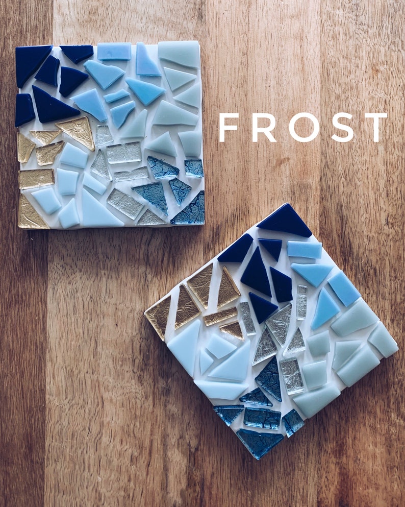 Puede incluir: Dos posavasos cuadrados con un dise&ntilde;o de mosaico en tonos azules, dorados y plateados. Los posavasos est&aacute;n hechos de vidrio y tienen un fondo blanco. La palabra "FROST" est&aacute; escrita en letras blancas sobre una superficie de madera.