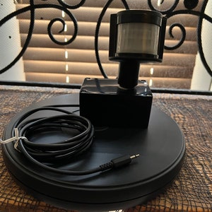 May include: A black motion sensor with a white sensor lens, a black box, and a black cord. The sensor is mounted on a black circular base.