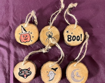May include: Six round wood ornaments with Halloween themes. The ornaments are painted with a black spider, a ghost, a pumpkin, a black cat, a bat, and the word "Boo!"