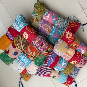 May include: A patchwork cushion with a variety of colorful fabric patterns. The cushion is made of squares of fabric stitched together, featuring floral, geometric, and abstract designs in shades of red, blue, green, and purple. It has a decorative tie.