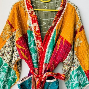 May include: A patchwork robe with a vibrant mix of colors, including yellow, red, green, and teal. The robe features floral and geometric patterns, with a red tie belt. The garment is hanging on a yellow hanger.