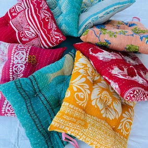 May include: A collection of colorful, handmade floor cushions. The cushions feature various patterns and colors, including red, turquoise, pink, and yellow. Each cushion has a unique design, with floral and geometric patterns. Some cushions have a button in the center.