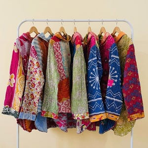 May include: A row of seven long-sleeved button-down shirts hanging on wooden hangers on a white metal rack. Each shirt features a unique patchwork design with vibrant, multi-coloured floral and geometric patterns in shades of pink, blue, green, and yellow.