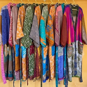 May include: A collection of colorful, patterned kimonos hanging on a clothing rack. The kimonos feature various designs in shades of blue, orange, green, and pink. The background is a solid yellow wall.