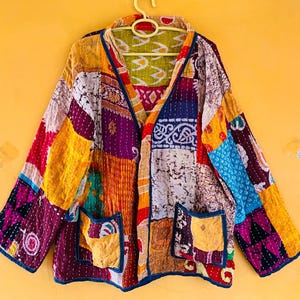 Puede incluir: Una chaqueta de manga larga con un diseño de patchwork que presenta varios patrones de tela coloridos en tonos amarillo, morado, azul y blanco. La prenda incluye dos bolsillos delanteros, un cuello en V y costuras blancas visibles.