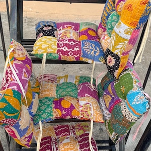 May include: A collection of colorful, patchwork cushions with various patterns and designs. The cushions are arranged on a black metal frame, showcasing a mix of vibrant colors like yellow, pink, green, and purple. Each cushion appears to be handmade.