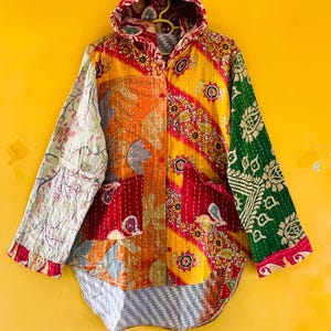 Puede incluir: Una chaqueta con capucha que presenta un diseño de patchwork con varios patrones florales y geométricos en tonos vibrantes de amarillo, naranja, rojo y verde. La prenda incluye mangas largas, cierre frontal de botones y dos bolsillos grandes.