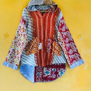 Op de afbeelding: Een jack met capuchon en lange mouwen met een patchworkontwerp met diverse bloemen- en geometrische patronen in tinten rood, oranje, blauw en wit. Het kledingstuk is voorzien van een knoopsluiting, twee voorzakken en een afgeronde zoom.